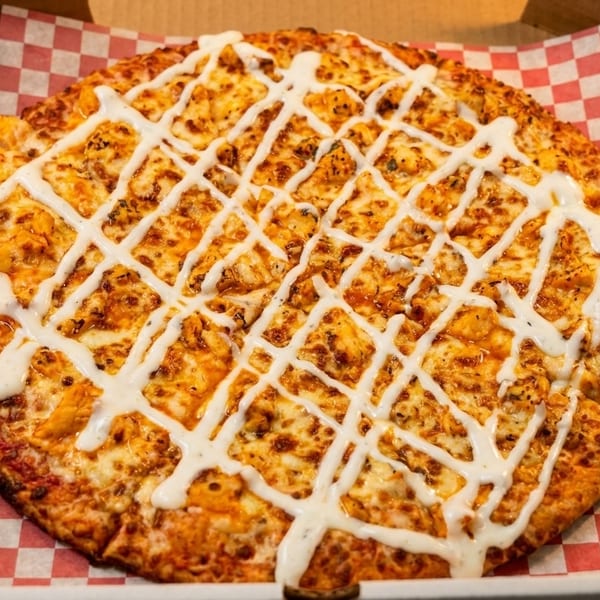 A buffalo chicken pizza with ranch drizzled on top, in a pizza box with red and white checkered paper.