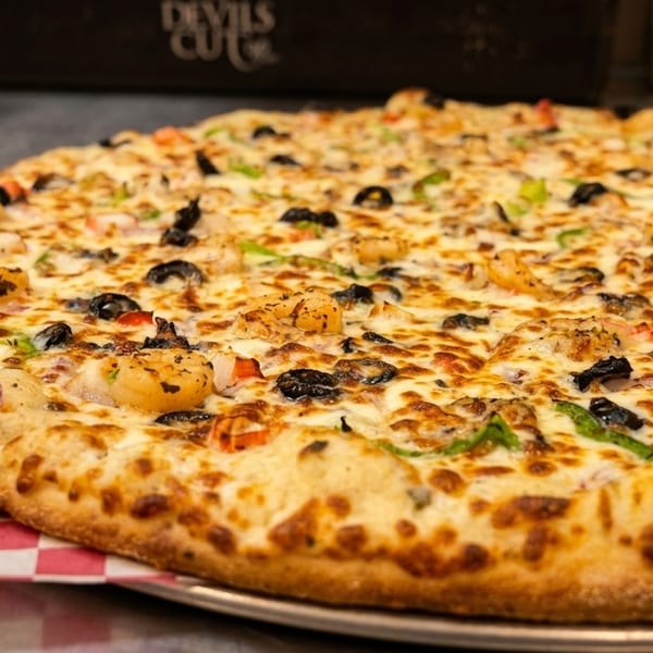 A large, freshly baked pizza with shrimp, black olives, and green peppers on a checkered paper.